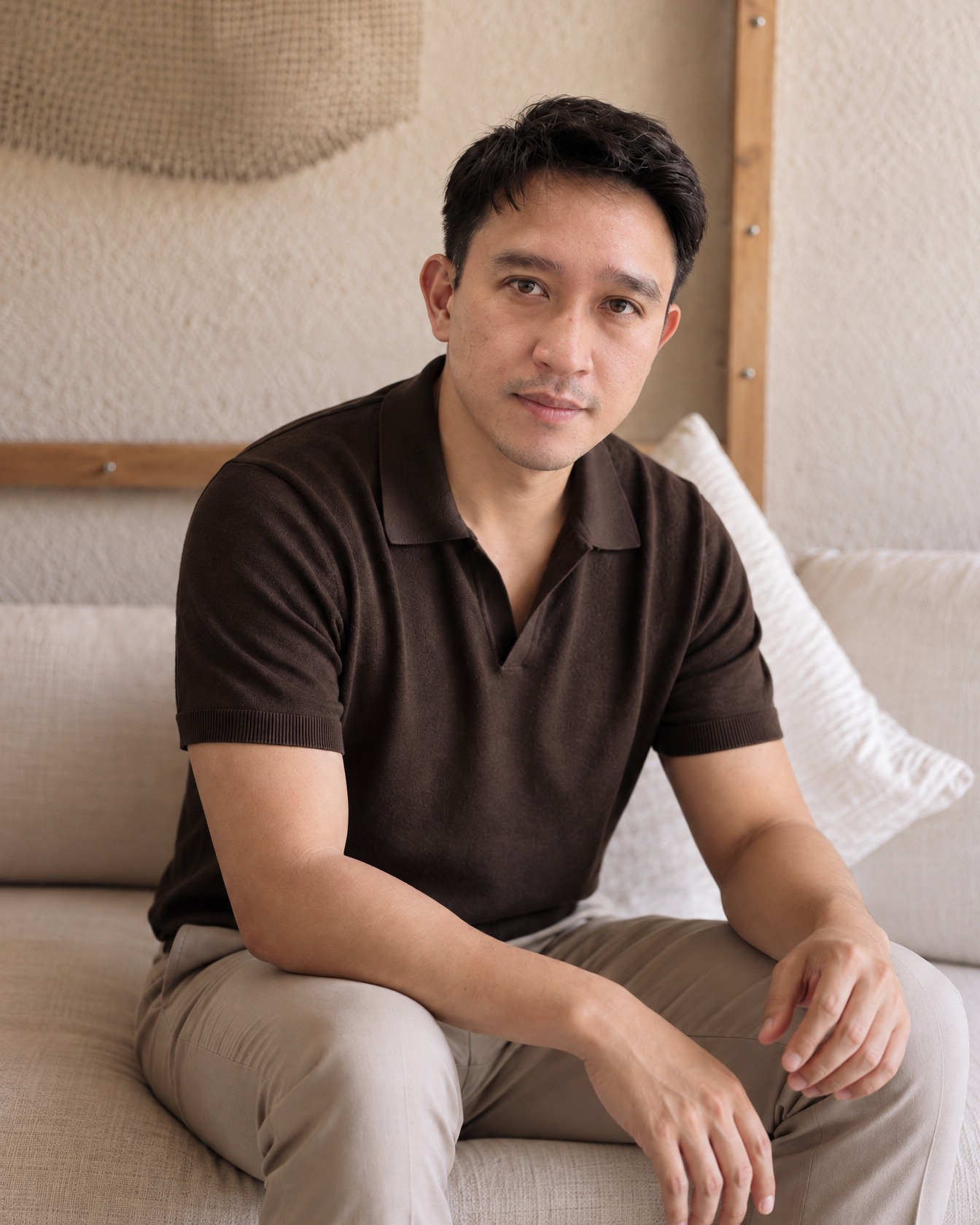 Nat Wongsaroj seated on a cream sofa in a brown polo shirt