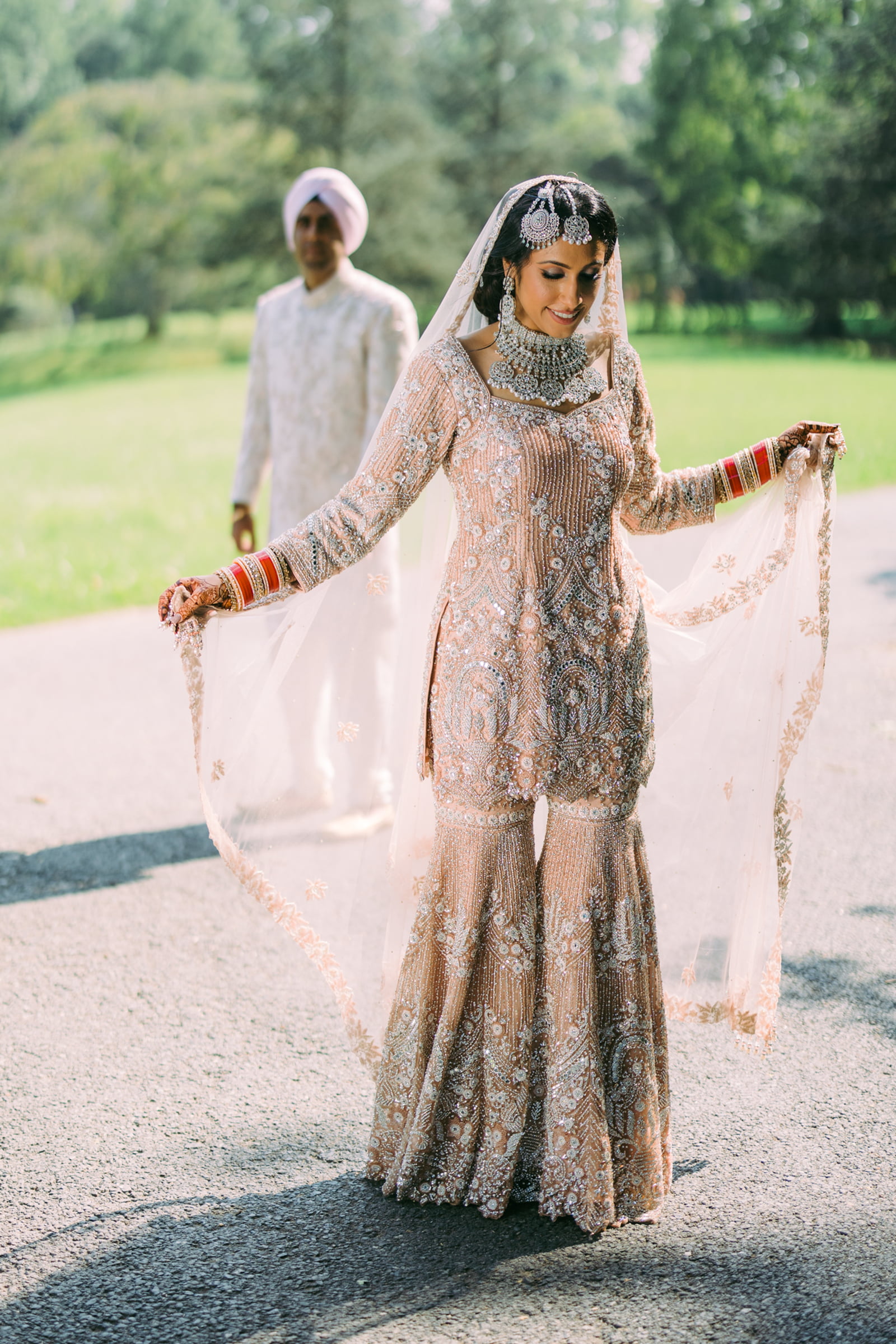 A bride holding her embroidered veil with the groom waiting behind her